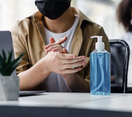 Focus on antiseptic to protect against covid virus. Guy disinfects his hands, sitting at table at workplace with laptop, working in interior of modern office, panorama, copy space